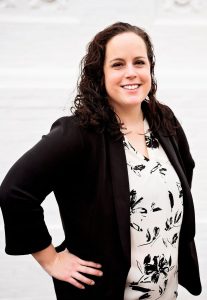 A headshot of a Lindsey Baker. She is a white woman with brown, curly hair past her shoulders. She wears a white button-down shirt with black flowers on it and a black blazer. Her hands are on her hips. The background is white.