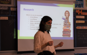 Lia Atanat, a white brunette woman, presents and stands in front of a prokector of a PowerPoint slide that says "Research." She is in a classroom and wears a mask, white sweater. The photo only goes down to her waist..