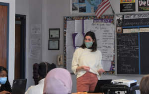 Lia Atanat, a white brunette woman, presents to students in a classroom. She wears a white sweater, pinkish pants and a mask. We see the back of heads of 3 students and the side of one's face. Behind her is a chalkboard and other classroom presentation materials.