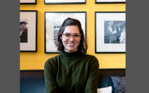 Headshot of Lia Atanat, a white bruette woman with shoulder-length hair and glasses. She wears a green turtleneck . She sits in an office on a black couch in front of a yellow wall with black-and-white photographs.