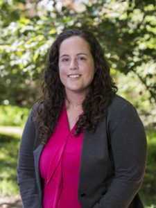 A headshot of a white woman with brown, curly hair past her shoulders wears a pink blouse and dark gray cardigan. In the background, slightly blurry, is a tree, grass, and pathway, and a bit of sky.