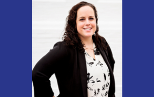 A headshot of a Lindsey Baker. She is a white woman with brown, curly hair past her shoulders. She wears a white button-down shirt with black flowers on it and a black blazer. Her hands are on her hips. The background is white.