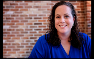 A headshot of a Lindsey Baker. She is a white woman with brown, curly hair past her shoulders. She wears a royal blue blouse with a V-neck and small dangly earrings. She is inside a building with brick background.