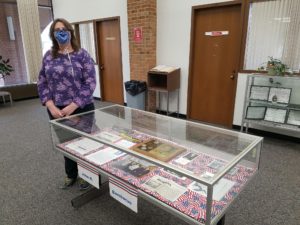 Yelizaveta L Zakharova, a white woman with layered brown hair, stands in a library to the left of an exhibit case. She wears a patterned purple shirt and black slacks. Next to her, an exhibit case displays pictures and documents of and about John H. Bambacus.