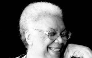 a Black and white photograph of Lucille Clifton from the chin up, in front of a black background. The photo is later in life and she is wearing glasses and laughing. Her hand rests near her chin.