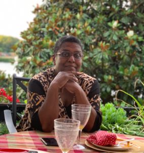 A Black woman with short hair sits behind a table covered in red checkered tablecloth and empty drink glasses. She sits in front of a tree with water in view behind her.