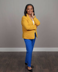 A Black woman standing on a wooden floor wearing a yellow blazer and royal blue pants.