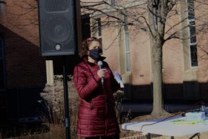 Congresswoman Odette Ramos in a burgundy winter coat standing with a microphone, wearing a mask. She stands outside in front of a speaker, with a tree in view.