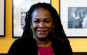 The late Marilyn Hatza, a middle-aged Black woman, sits for a professional portait. She has her in locs and down, wears a magenta printed dress with a black sweater, along with a colorful necklace and matching bracelet. She sits front of a yellow wall with black and white photographs.