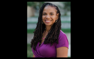 A professional photograph of Dr. Kimberly R. Moffitt, a Black woman smiling. She has dreadlocks in a side part. She wears a short-sleeved magenta blouse and is in front of a blurry outdoor setting.