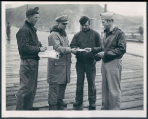 A black and white image of four World War II Coast Guardsmen and soldiers receive their voting applications while stationed in the Aleutian Islands. To their left, a man in the background looks on behind them. To their right, there is an airplane.