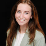 A headshot of white women with auburn hair: she wears a gray blazer, white shirt, and a gold necklace. Her hair is down and the background is black