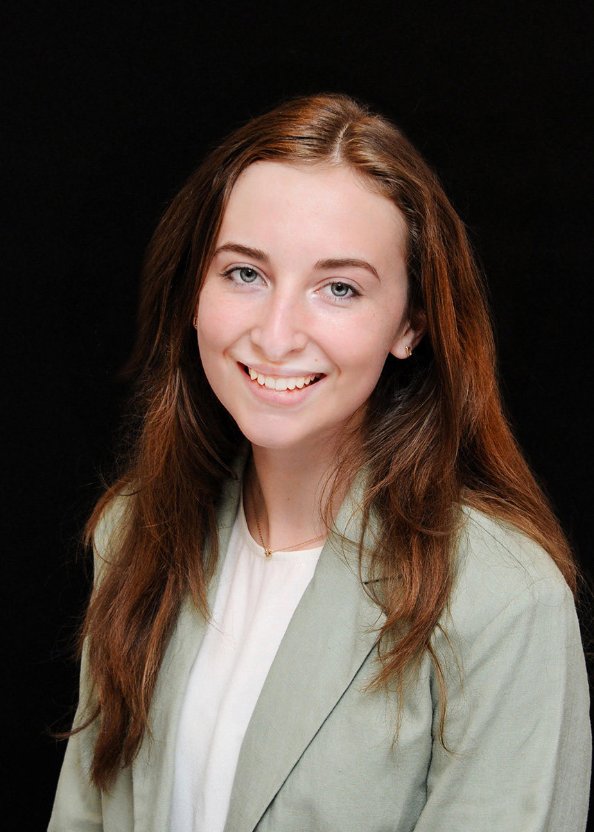 A headshot of white women with auburn hair: she wears a gray blazer, white shirt, and a gold necklace. Her hair is down and the background is black