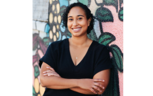 Naima Coster, an Afro-Latina woman with black curly hair down to about her chin. She smiles and wears a short sleeved, black shirt, a silver star, and small stud earrings. (The photo is to about her waist.)She stands with her arms crossed, not unfriendly, in front of a colorful mural.