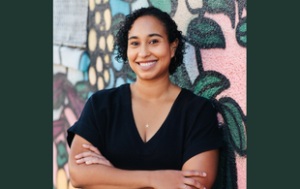 Naima Coster, Afro-Latina woman with black curly hair down to about her chin. She smiles and wears a short sleeved, black shirt and jeans. She also wears a silver necklace of a star and small stud earrings. She stands with her arms crossed in a professional manner and stands in front of a colorful mural.