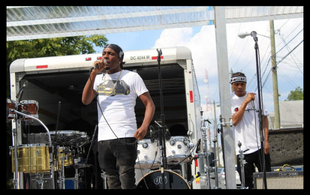 An outdoor music festival. 2 Black male musicians wearing white t-shirts and black pants. They are in front of an intricate drum set-up onstage. We see the back of the white truck or van, where it looks like the drums were loaded. It is a sunny day, with blue sky and some clouds.