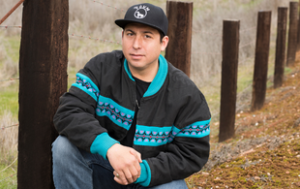 professional photograph of Tommy Orange, a member of the Cheyenne and Arapaho Tribes of Oklahoma. He wears a patterned jacket, black shirt, blue jeans, and baseball cap and sort of sits/crouches down. He is outside, surrounded by trees.