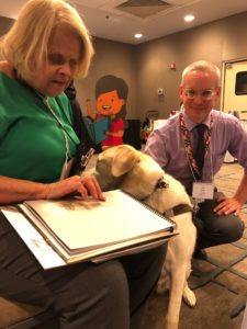 A library patron, a white middle-aged woman with a teal short-sleeved shirt, sits with her braille book and her service dog, which looks like a retriever. John Owen, a white man wtih gray hair and glasses, wearing a purple button-down shirt and khakis, sits next to her and smiles for the camera.