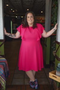 Lindsey Baker, a white woman in her thirties, stands inside and smiles. She has curly brown hair, wears a knee-length bright pink dress, and colorful sneakers. She also has various tattoos on both arms. Lindsey leans her arm on a wall/post next to her.The setting appears to be a greenhouse but could simply be a room with many plants in it.