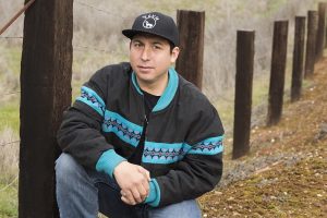 photo of Tommy Orange wearing jeans, a gray and turquoise jacket and a black baseball cap