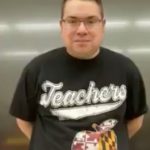 Joshua Pleasant, a white man, stands in front of a blackboard. He wears a black t-shirt with white print in the style of a baseball jersey that says "Teachers." Under the word is an apple with the pattern of the Maryland flag.