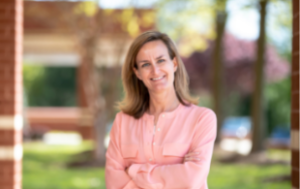 Lynn Rashid, a white woman with dirty blonde hair in a long-sleeved pale pink shirt stands outside in front of an intentionaly blurry background, with three trees To her left, we see a few inches of a brick building.