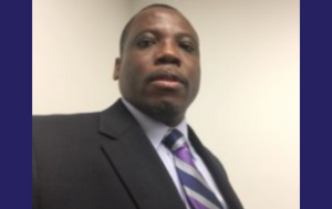 An image of of a Romuladus Azuine, a Black man. We see his head and the upper portion of his torso. He wears a Black suit jacket, a white button down shirt, and a tie with navy blue, purple, and light gray stripes. The background is off-white.