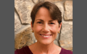 A headshot of Liz Cannon, a smiling white woman with cropped brown hair, from the shoulders up. Liz wears an eggplant-colored shirt with a scoop neck and small gold earrings. She is in front of a sandy-colored brick background.