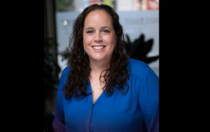 a professional headshot of Lindsey Baker. She is a white woman with brown, curly hair past her shoulders. She wears a royal blue blouse that fades into purple. The second image is a logo.