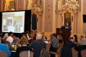 Image of Lindsey Baker, a white woman, giving a keynote presentation at a conference. On a screen, there are some photos from her life.