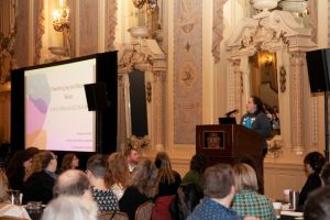 A white woman gives a keynote address at a conference. There is a screen with a slide that is the name of the presentation: "Prioritizing Joy and Showing Grace: A Small Museum Call to Action"