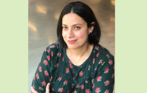 A professional headshot of Saima Sitwat, a Pakistani American woman. She has shoulder-length black hair and hazel eyes. She wears a green shirt with pink and yellow flowers on it. Her arms are crossed.