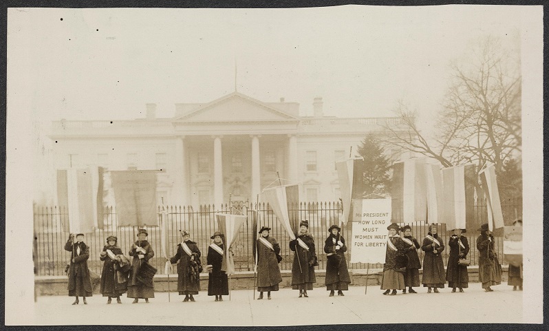 A 1917 black and white photograph of 13 white suffragettes in dark coats and sashes in front of the White House. Some have picket signs.