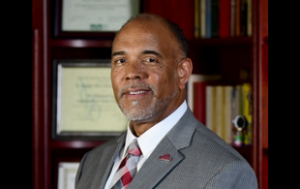 A professional photograph of Marshall F. Stevenson, Jr. He is an older Black man with a gray beard. He smiles and wears a gray sports jacket, a white button-down shirt and patterned tie with red, white, and gray. The background of the headshot is his office with books and his diploma.