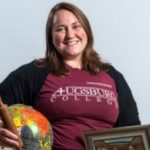 Laya Theberge, a white woman with light brown hair, wears a maroon t-shirt with "Augsburg College" on it and a black cardigan. She holds a globe and rolling pin in one hand an a framed painting in the other (the painting itself is not visible).