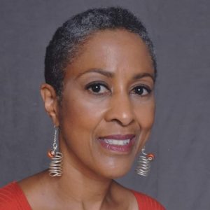 A Black woman with short, cropped hair wears dangly, silver earrings with orange stones. She wears and orange shirt. The image goes to the top of her shoulders.