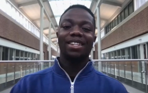 Augustus Roberts, a college student, He is Black with short hair and wears a royal blue zip-up sweater. He is in a school building.