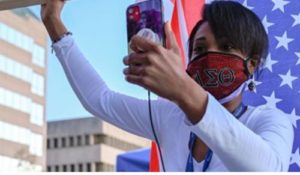 A Black woman in front of an American flag sitting vertically. She wears a white, long-sleeved shirt, red face mask, and raises her fist.