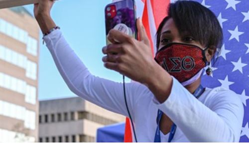 VVEEP Sun Image A Black woman in front of an American flag sitting vertically. She wears a white, long-sleeved shirt, red face mask, and raises her fist.