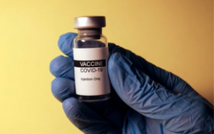 An image of a hand in a medical glove holding a clear vial marked "Vaccine COVID-19." Under the vaccine's name, the bottle says "Injection only." The label is white except the word "Vaccine" is on a small, black highlighted portion in white. The rest of the font is black.