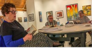 Three white adults, middle-aged to older, sit at a round table looking at their books. There is artwork of nature on the walls.