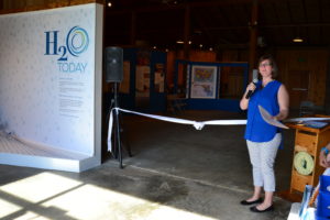 A sign that says "Maryland H2O" with Rachelle Green, a white woman with brown hair and glasses wearing a royal blue sleeveless button-downn, light capris, and black flats.