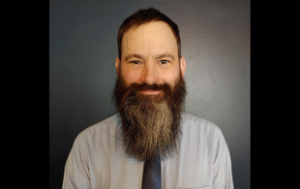 Michael Yuscavage, a white man with brown hair and full beard that goes several inches past his chin. He wears a gray button-down shirt and patterned tie. Michael smiles sits in front of a darker gray photography backdrop. 