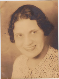 August Chissell in an old black-and-white photograph of her head and shoulders. She is a lighter-skinned Black woman with dark black hair. She smiles and looks to be wearing a cardigan. Due to the picture's age, it looks more light brown and black, than black and white.